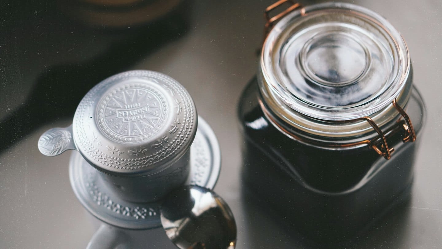 A metal coffee filter and glass jar on a surface.