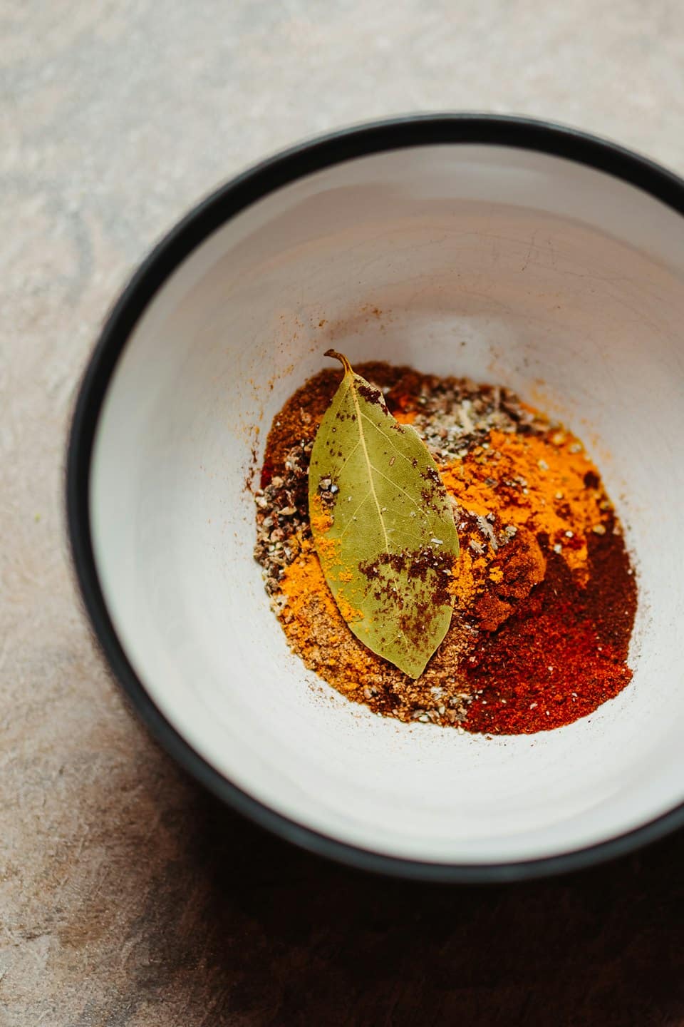 White bowl filled with colorful ground spices and a bay leaf.