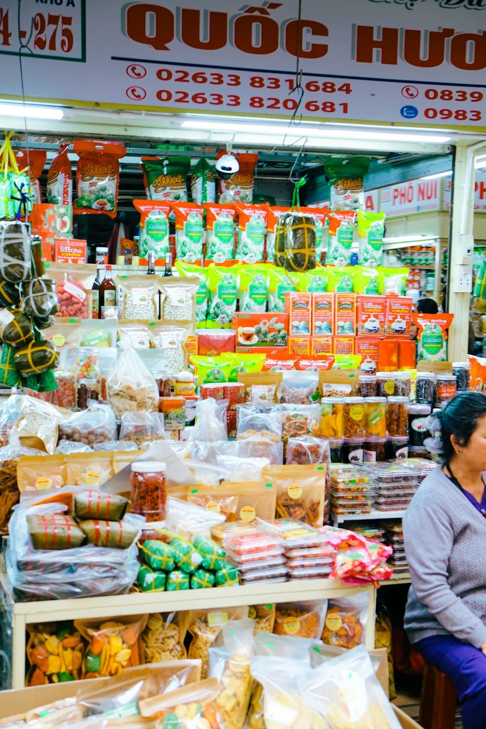 A market stall vendor sits among shelves filled with traditional food products.
